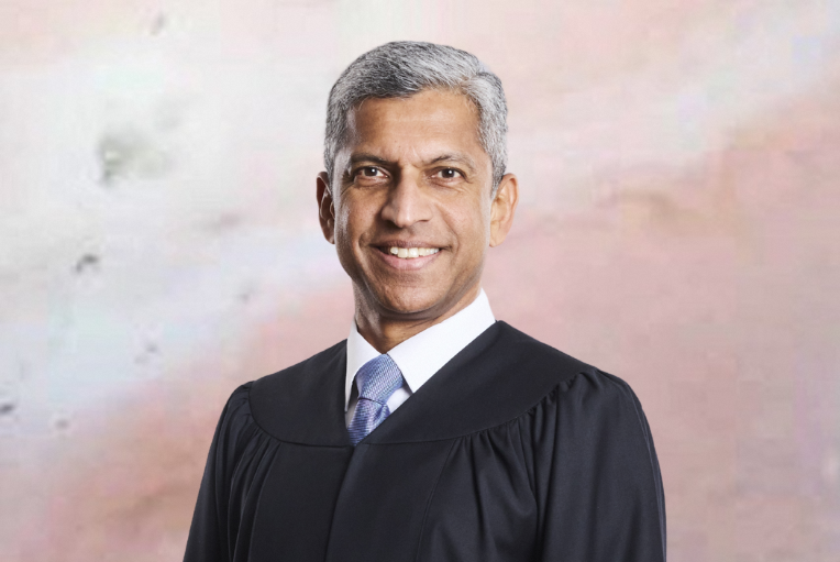 Man in judicial robe smiles; gray hair, white shirt, blue patterned tie, neutral background.